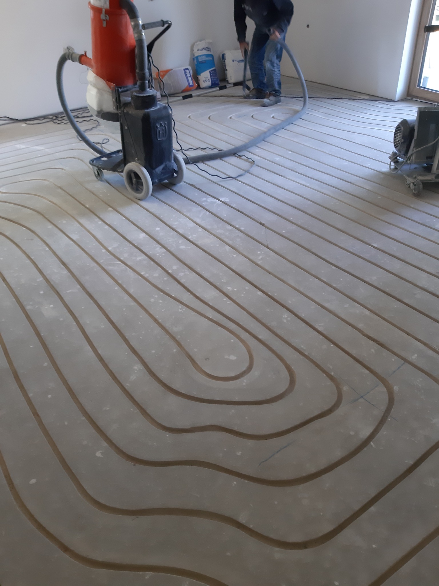 Widok z góry na rury instalacji grzewczej zatopione w wylewce, z widocznym odkurzaczem przemysłowym Husqvarna i pracownikiem w tle.