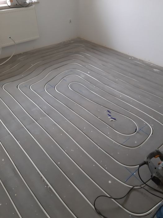 Białe rurki systemu rozprowadzone na szarej wylewce podłogowej, widoczne niebieskie znaczniki i fragment narzędzia elektrycznego w rogu pomieszczenia.