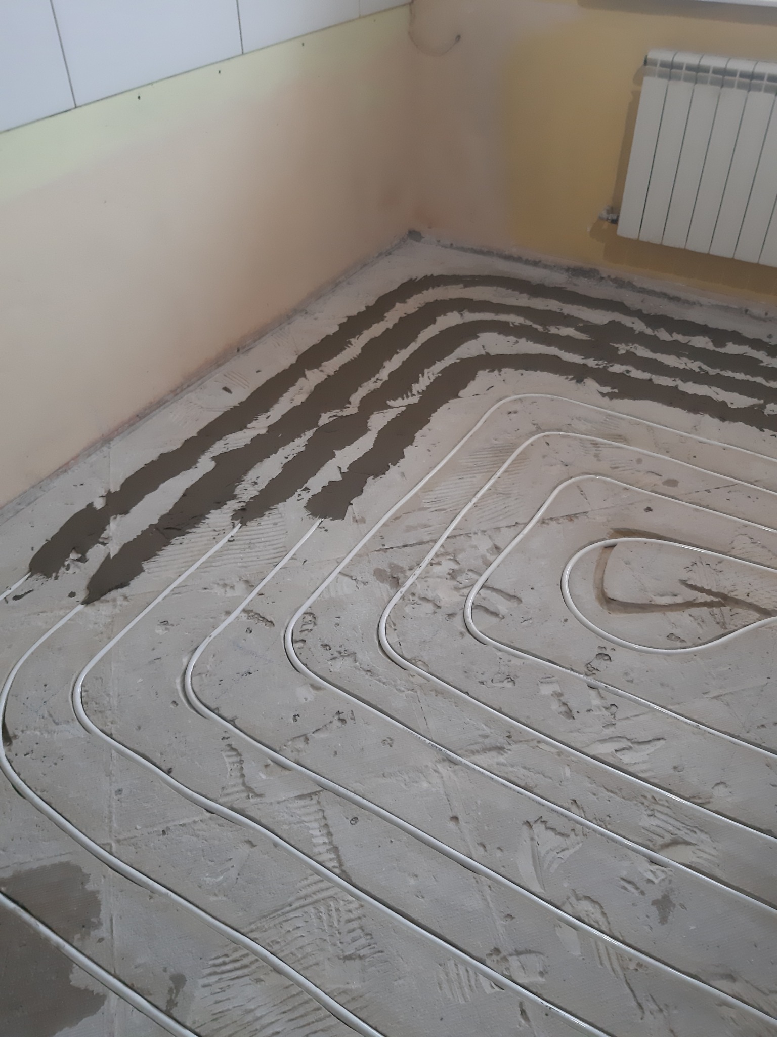 Białe rurki systemu grzewczego ułożone w betonowej wylewce podłogowej, widoczny grzejnik na ścianie.