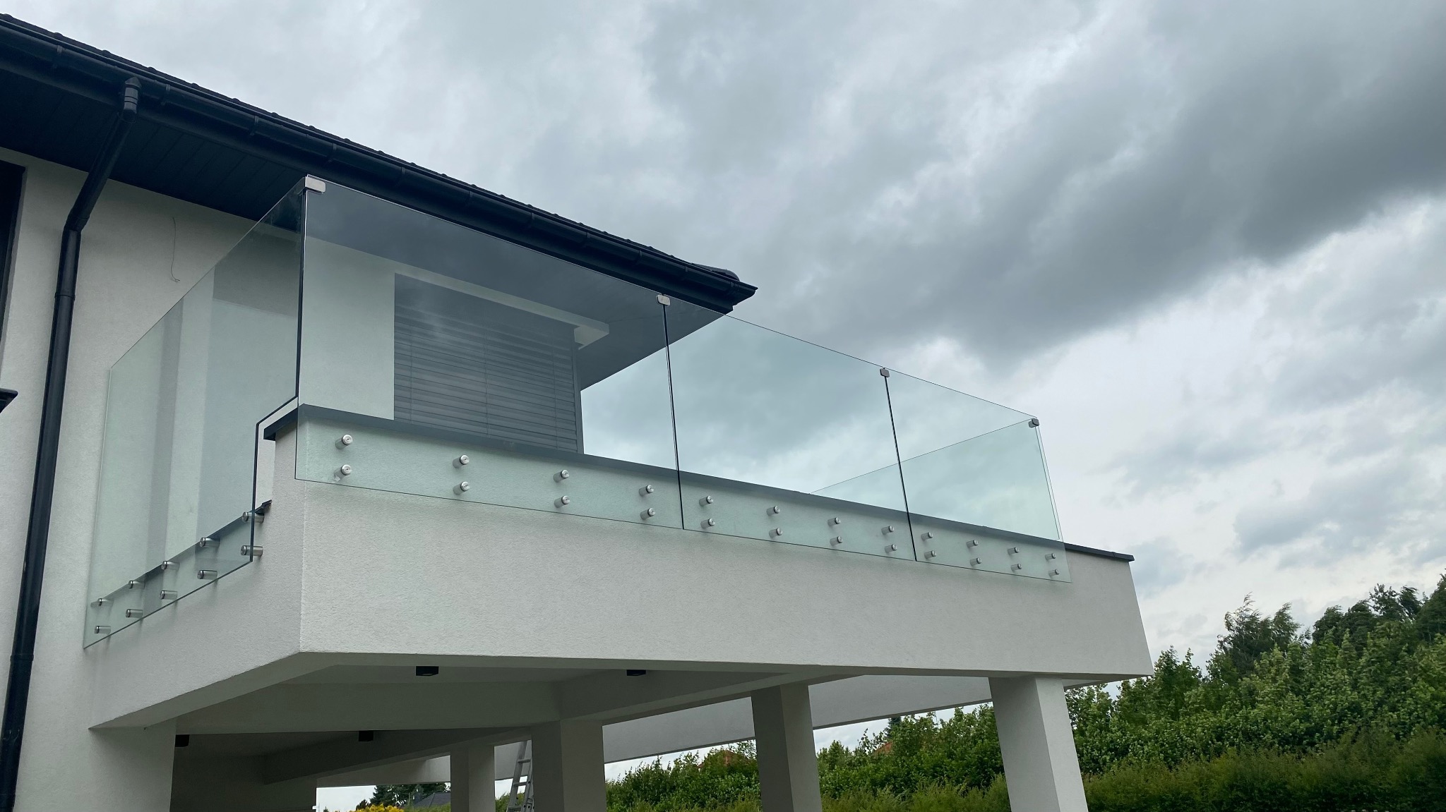 Szklana balustrada na balkonie nowoczesnego domu, mocowana punktowo do betonowej konstrukcji, widok z dołu na tle pochmurnego nieba.