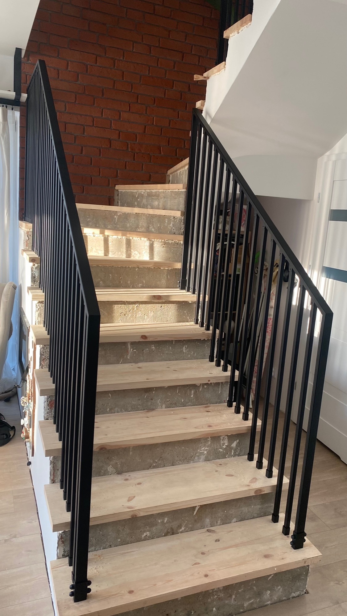 Wnętrze domu w trakcie remontu: surowe schody z drewnianymi stopniami i czarną, metalową balustradą na tle ściany z czerwonej cegły.