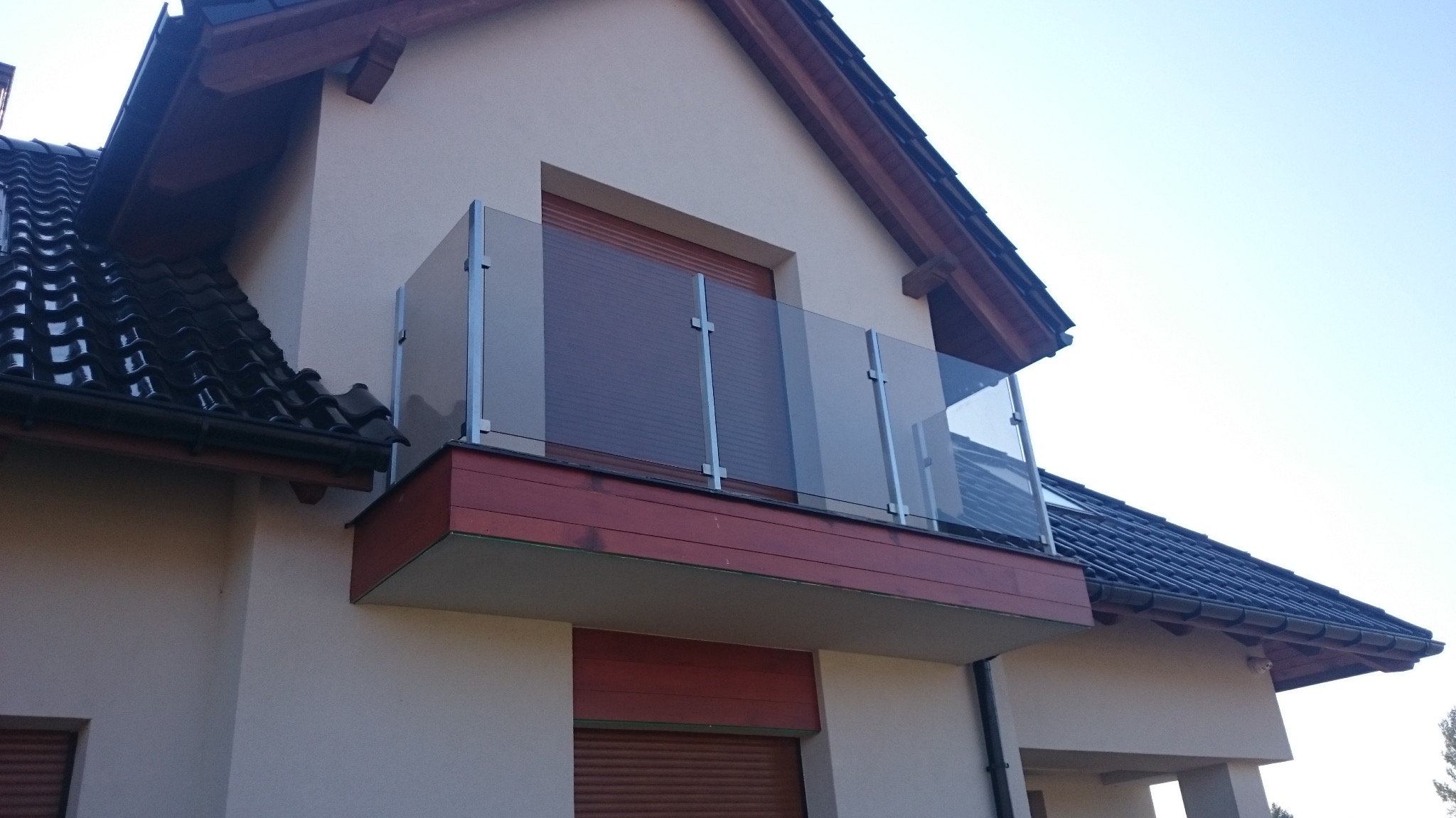Zewnętrzny balkon z nowoczesną szklaną balustradą, wspartą na metalowych słupkach, zamontowany przy domu z ciemną dachówką.