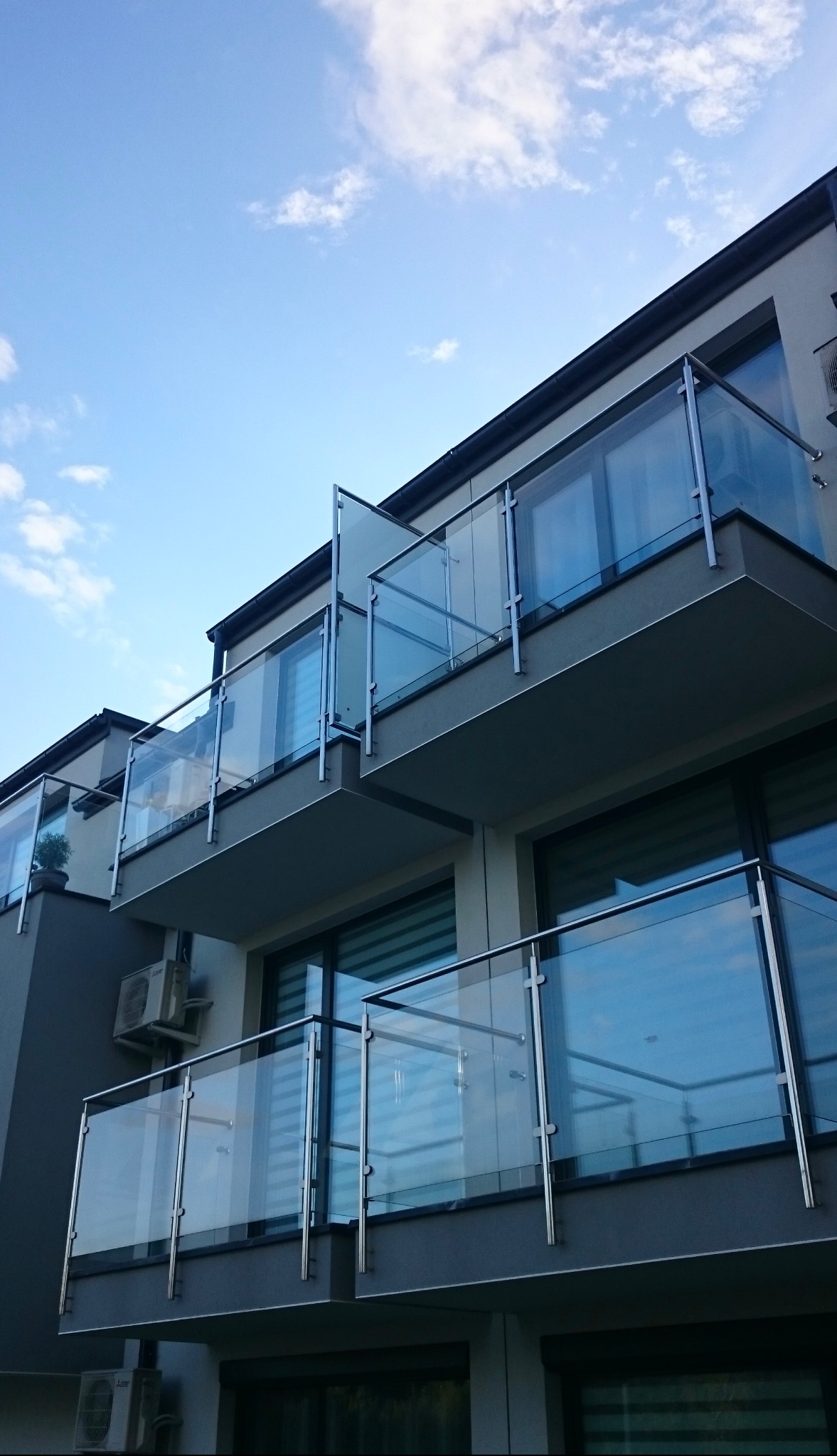 Nowoczesne balkony z przezroczystymi szklanymi panelami i metalowymi poręczami, widok z dołu na tle błękitnego nieba.