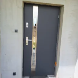 Szare drzwi zewnętrzne z pionowym, metalowym akcentem w Oleśnicy, widoczne podczas montażu w surowym otoczeniu, z wyeksponowaną ościeżnicą i fragmentem elewacji w trakcie wykończenia.