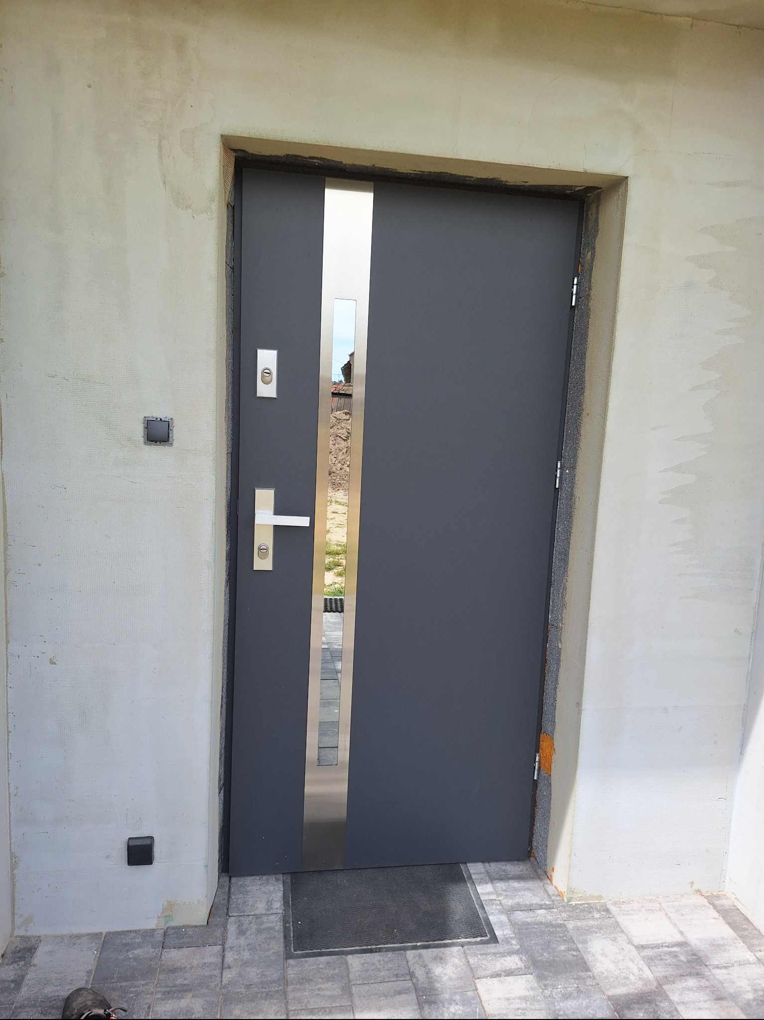 Szare drzwi zewnętrzne z pionowym, metalowym akcentem w Oleśnicy, widoczne podczas montażu w surowym otoczeniu, z wyeksponowaną ościeżnicą i fragmentem elewacji w trakcie wykończenia.
