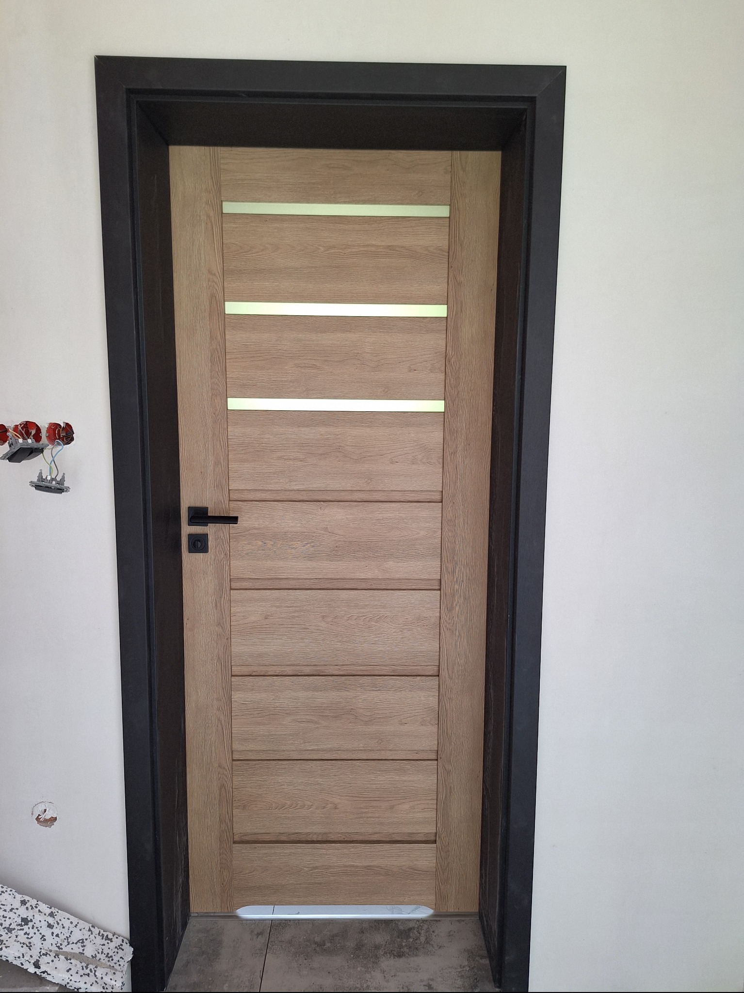 Zamknięte, dębowe drzwi wewnętrzne z poziomymi, srebrnymi intarsjami, czarną klamką i ościeżnicą, widoczne gniazdko elektryczne w ścianie obok.