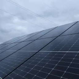 Panel słoneczny ustawiony pod kątem na tle pochmurnego nieba z widocznymi liniami energetycznymi w oddali.