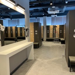 Patrycja Sutryk Czysty Kącik - Wnętrze szatni z rzędem szafek z numerami, lustrem z suszarkami do włosów i metalową drabinką w tle.