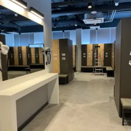 Wnętrze szatni z rzędem szafek z numerami, lustrem z suszarkami do włosów i metalową drabinką w tle.