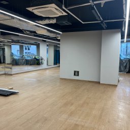 Patrycja Sutryk Czysty Kącik - Pusta sala z panelami podłogowymi w odcieniach jasnego drewna, duże lustra na ścianie, widoczne elementy instalacji pod sufitem, okna zabezpieczone folią ochronną, w tle sprzęt sportowy przykryty...