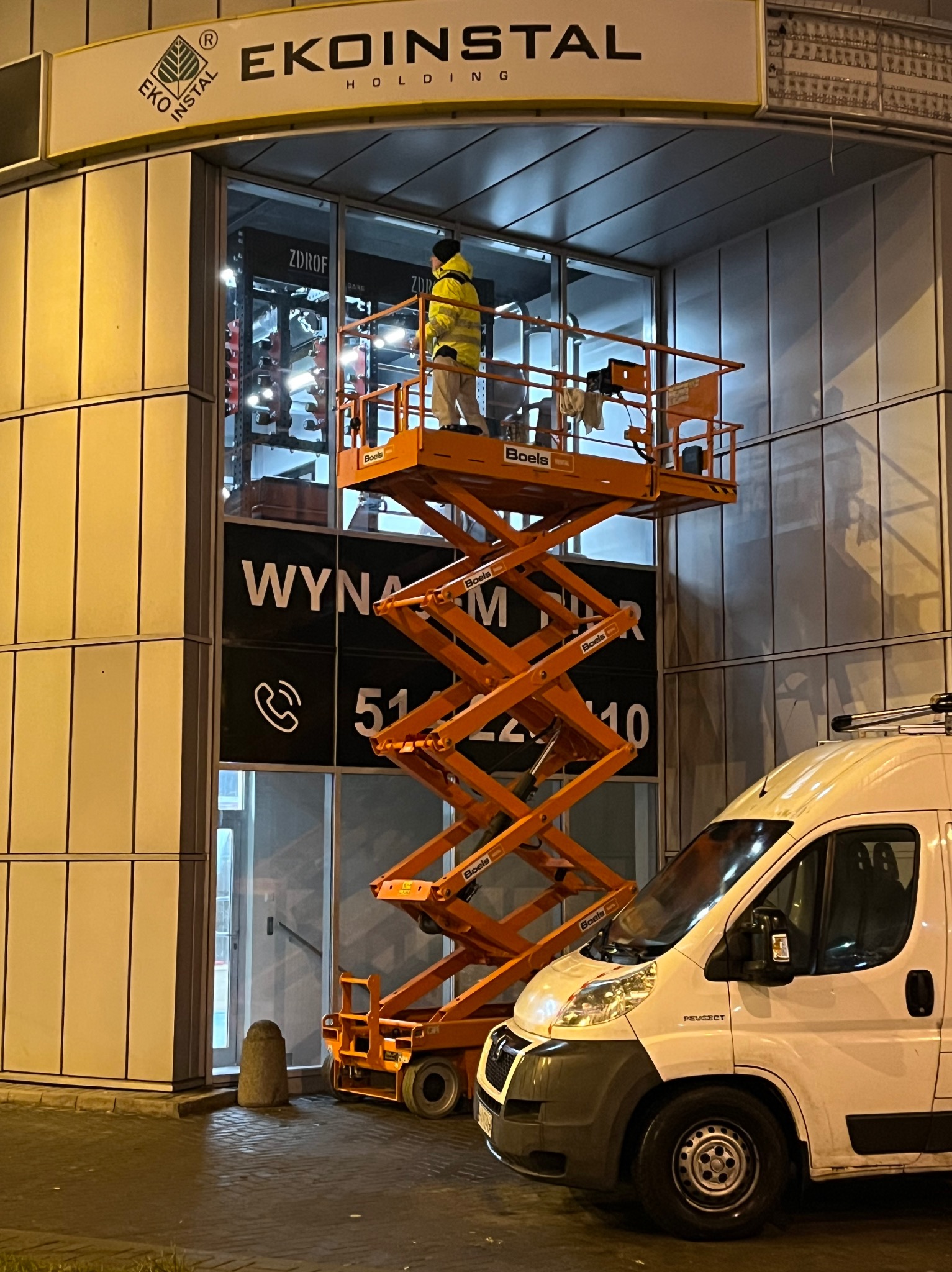 Pracownik w żółtej kurtce na podnośniku koszowym myje okna budynku z logo 'EKOINSTAL HOLDING', widoczny numer telefonu na oknie i zaparkowany biały van.