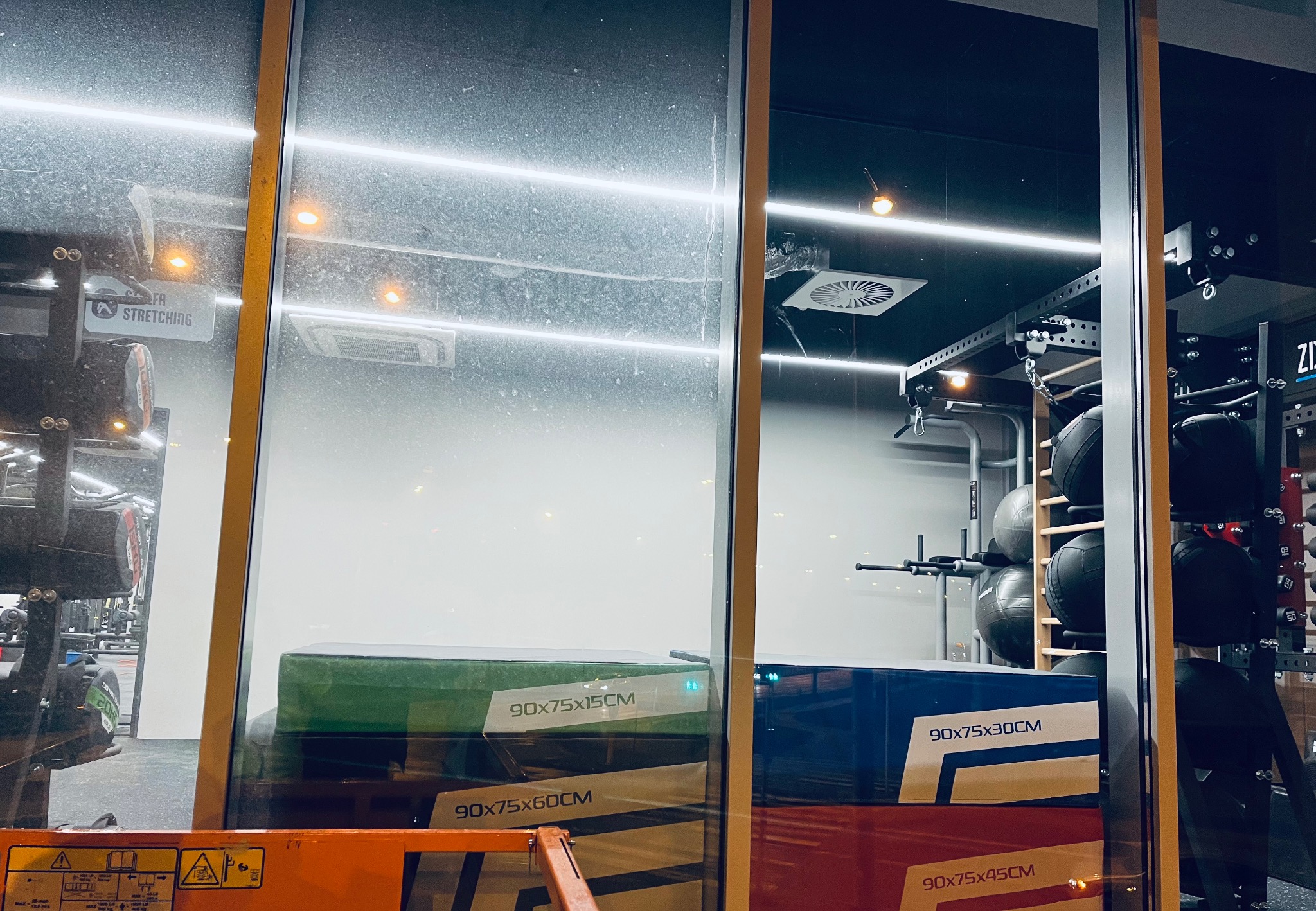 Brudna szyba okienna z widoczną refleksją wnętrza siłowni, na pierwszym planie ustawione kartony z akcesoriami do ćwiczeń.