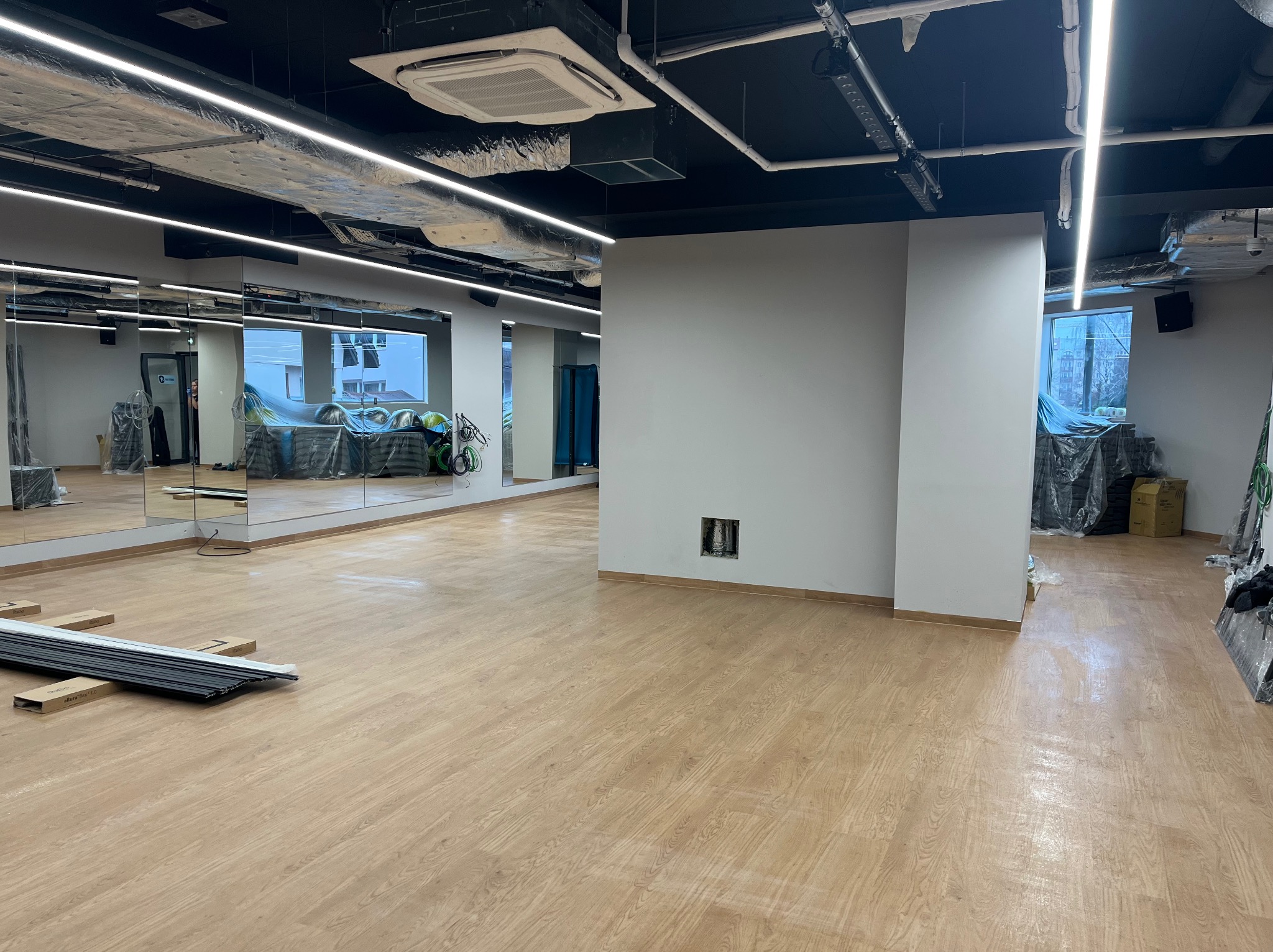 Pusta sala z panelami podłogowymi w odcieniach jasnego drewna, duże lustra na ścianie, widoczne elementy instalacji pod sufitem, okna zabezpieczone folią ochronną, w tle sprzęt sportowy przykryty...