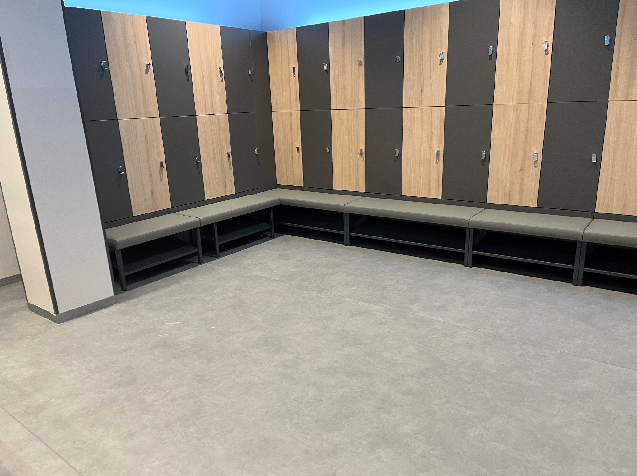 Wnętrze szatni z rzędem zamykanych szafek w kolorach drewna i grafitu, oraz szarą ławką z metalową konstrukcją.