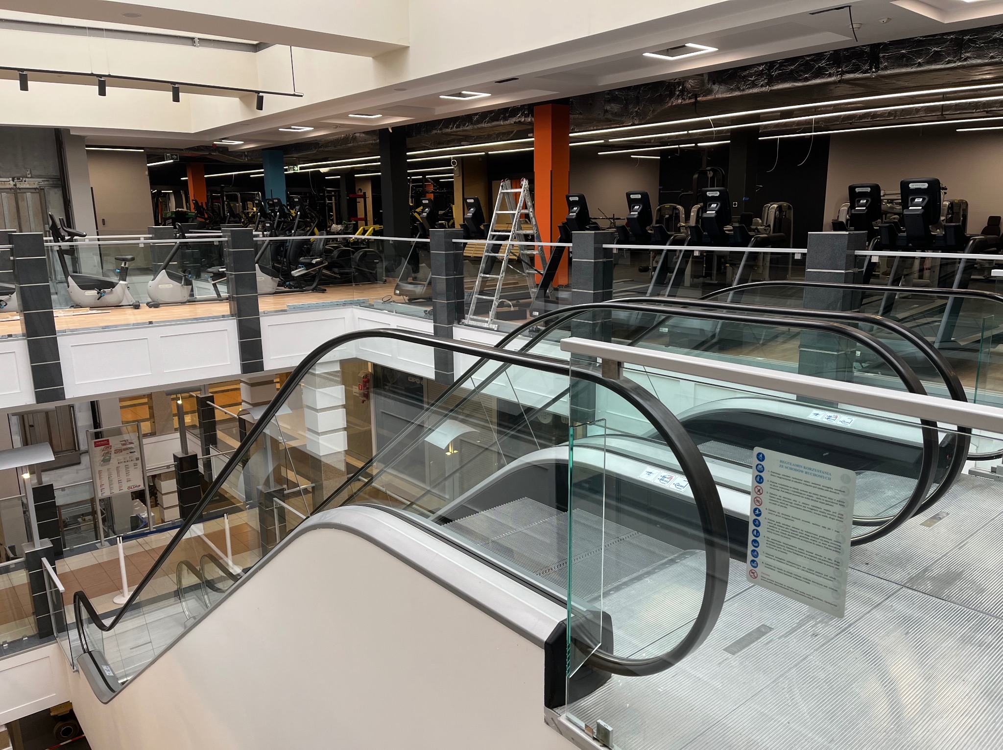 Wnętrze dwupoziomowego fitness klubu z widokiem z góry na schody ruchome i sprzęt do ćwiczeń na wyższym poziomie, drabina oparta o kolumnę.