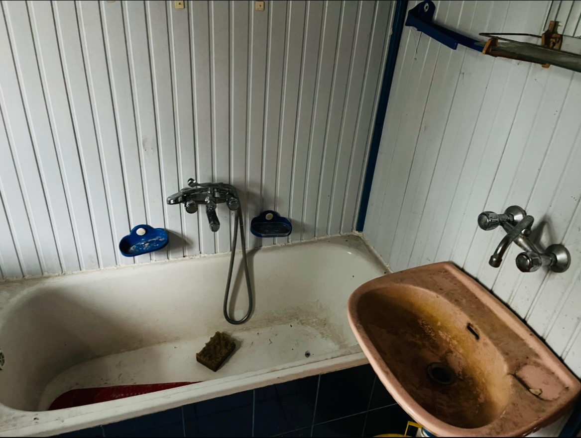 Zaniedbana łazienka z brudną wanną, umywalką i ścianami z paneli PCV, widoczna gąbka w wannie.
