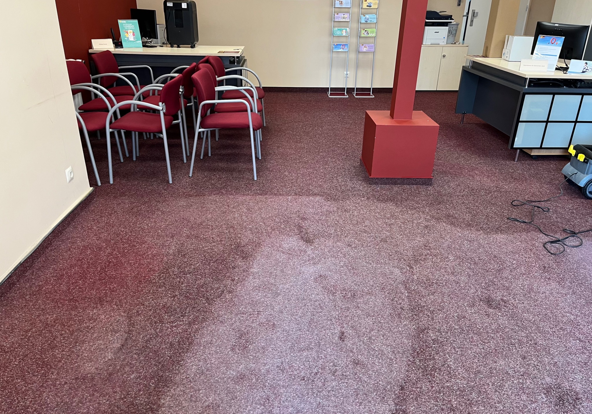 Częściowo wyczyszczony, bordowy dywan w biurze z krzesłami i biurkami, widoczne mokre plamy po praniu.