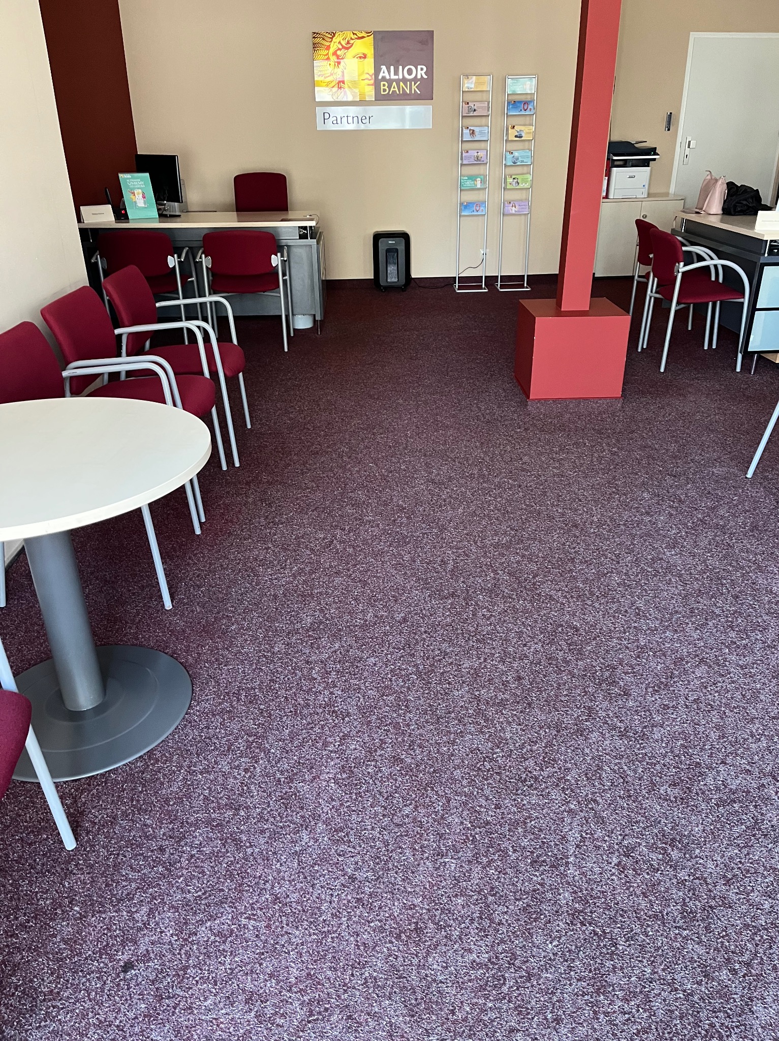 Wnętrze biura z ciemnoczerwoną wykładziną dywanową, białym okrągłym stołem i czerwonymi krzesłami, widoczne stanowiska pracy i materiały reklamowe Alior Bank.