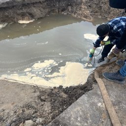 PG Construction Patryk Gul - Pracownicy w trakcie czyszczenia wykopu wypełnionego wodą z białym osadem, używając pompy zanurzeniowej i węża. Widoczny fragment drogi z uszkodzoną nawierzchnią.