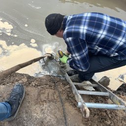 PG Construction Patryk Gul - Pracownik w niebieskiej koszuli w kratę i czapce pracuje przy pompie zanurzonej w zbiorniku z brudną wodą, stojąc na aluminiowej drabinie.