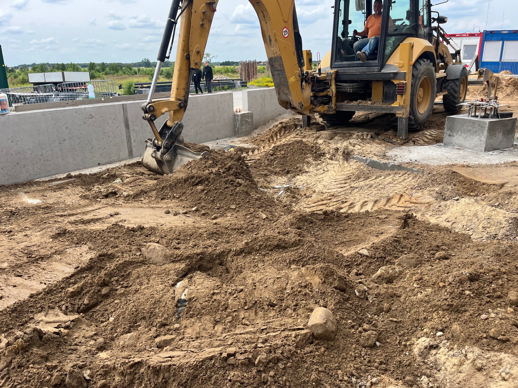 Żółta koparka przygotowuje teren pod fundamenty domu z betonowych prefabrykatów, widoczny operator w kabinie i dwóch mężczyzn w tle.