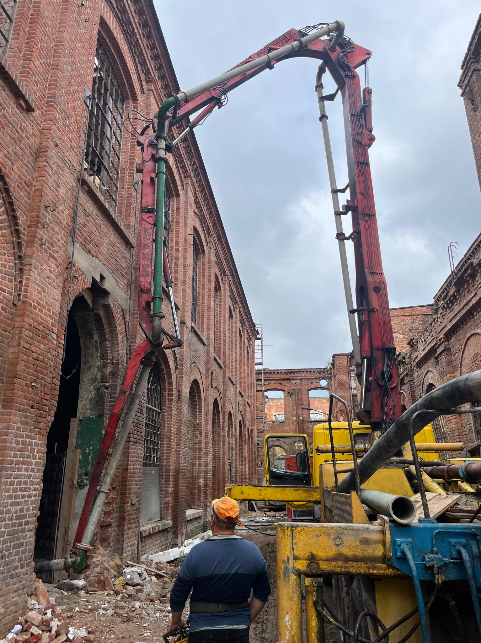 Czerwony wysięgnik pompy do betonu oparty o ceglany budynek w trakcie renowacji; widoczny pracownik w pomarańczowej czapce oraz żółty pojazd budowlany.