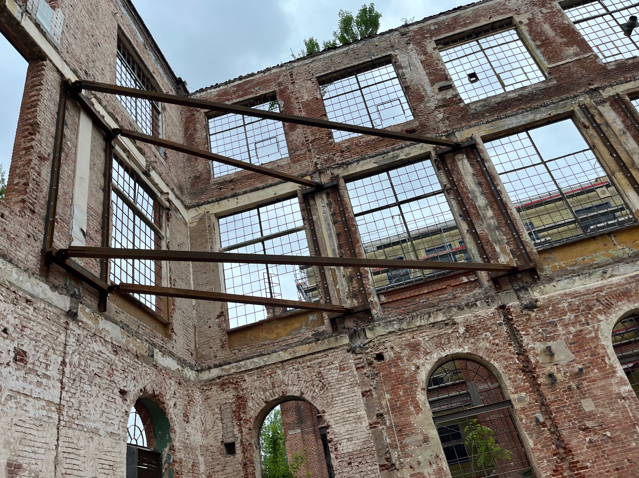 Opuszczona, ceglana konstrukcja budynku w Łodzi z licznymi oknami bez szyb i z widocznymi elementami metalowymi. W tle widoczny inny budynek.