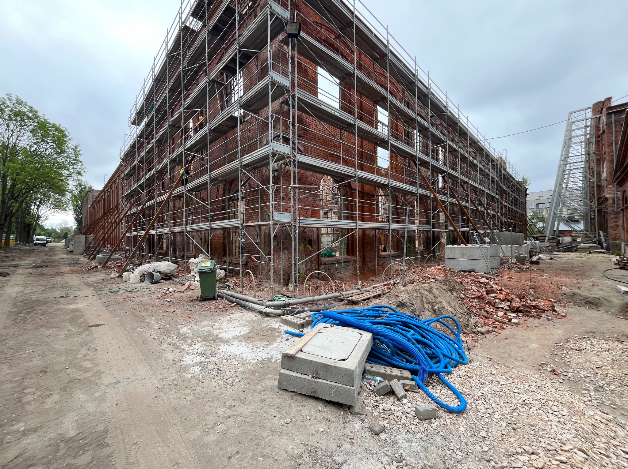 Budynek z czerwonej cegły w trakcie renowacji, otoczony rusztowaniami, z widocznymi niebieskimi rurami i gruzem na pierwszym planie, oraz pracownikami na rusztowaniach.