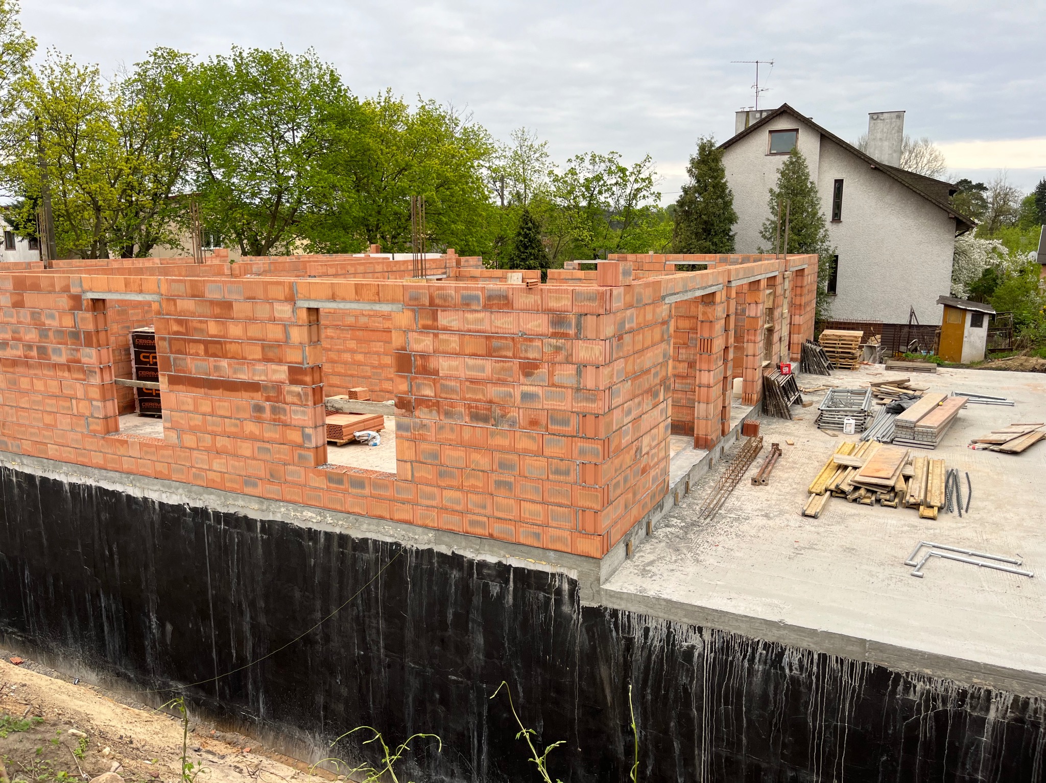 Wznoszenie ścian parteru domu z czerwonej cegły ceramicznej, widoczna izolacja fundamentów, w tle dom jednorodzinny i drzewa w wiosennej zieleni.