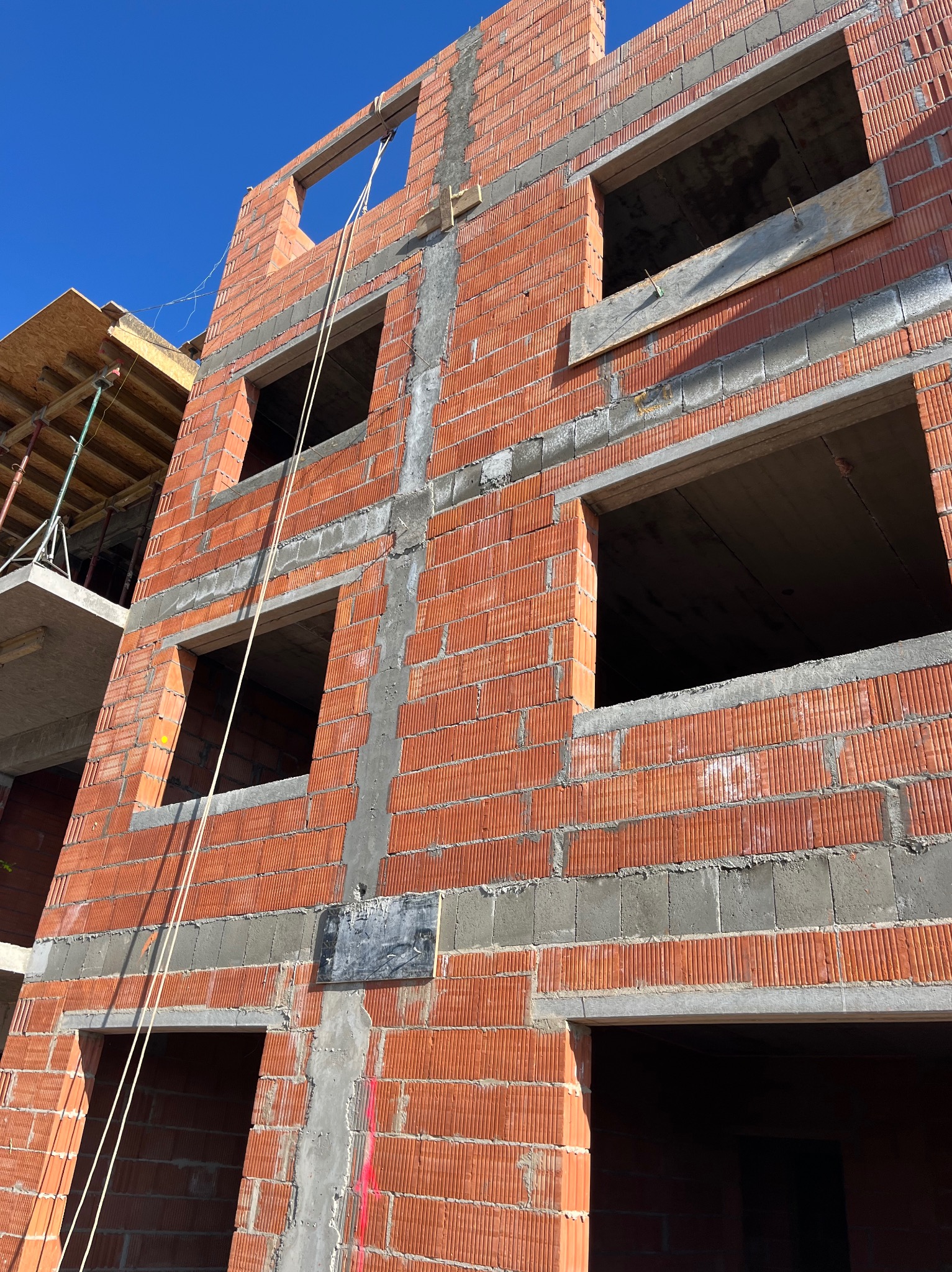 Wznoszona ściana budynku z czerwonej cegły ceramicznej z widocznymi otworami okiennymi i fragmentem rusztowania z boku, w słoneczny dzień.