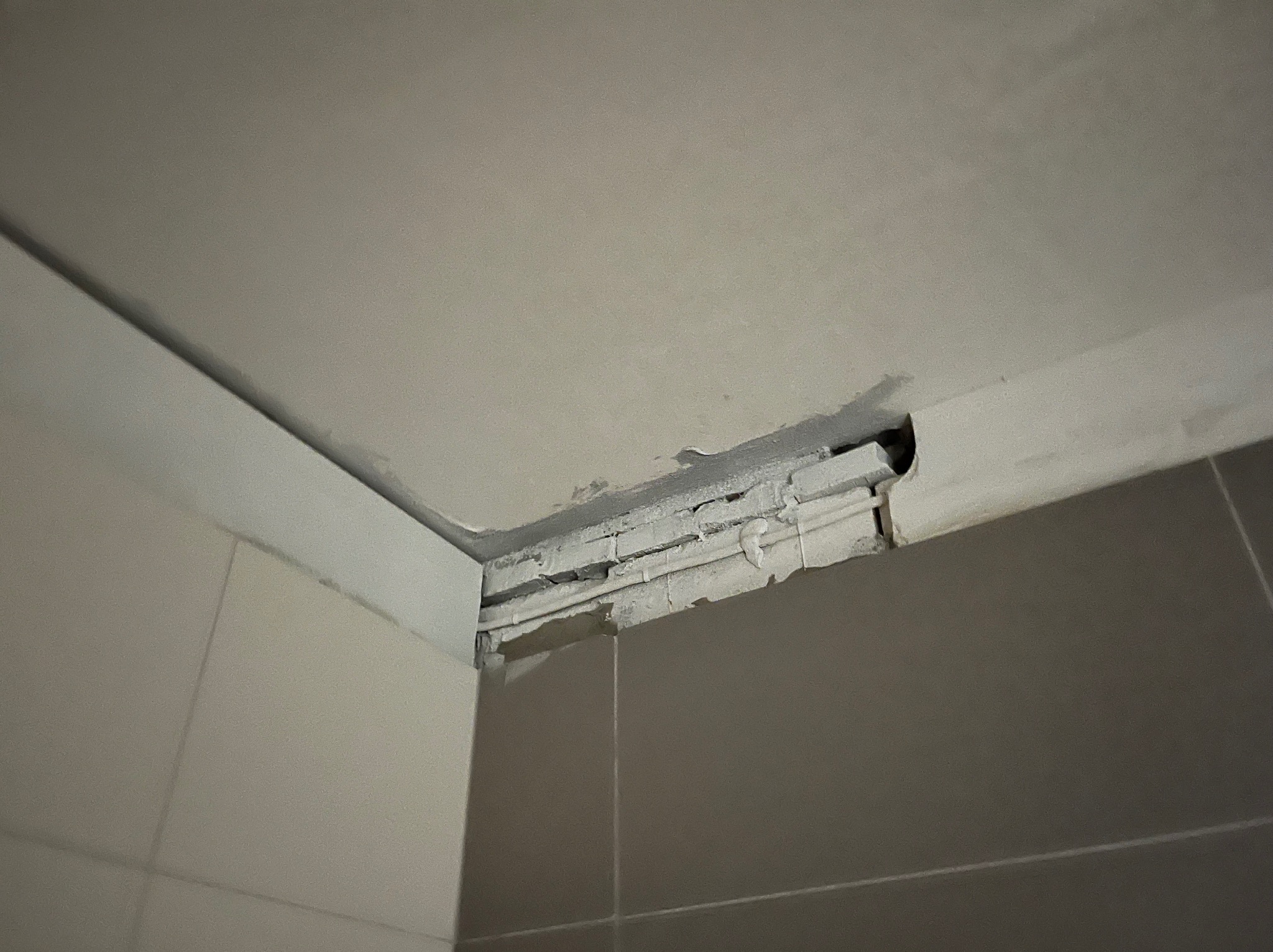 Niedokończone połączenie ściany z sufitem w łazience, widoczne rury i niedokładności w wykończeniu, szare płytki na ścianie.