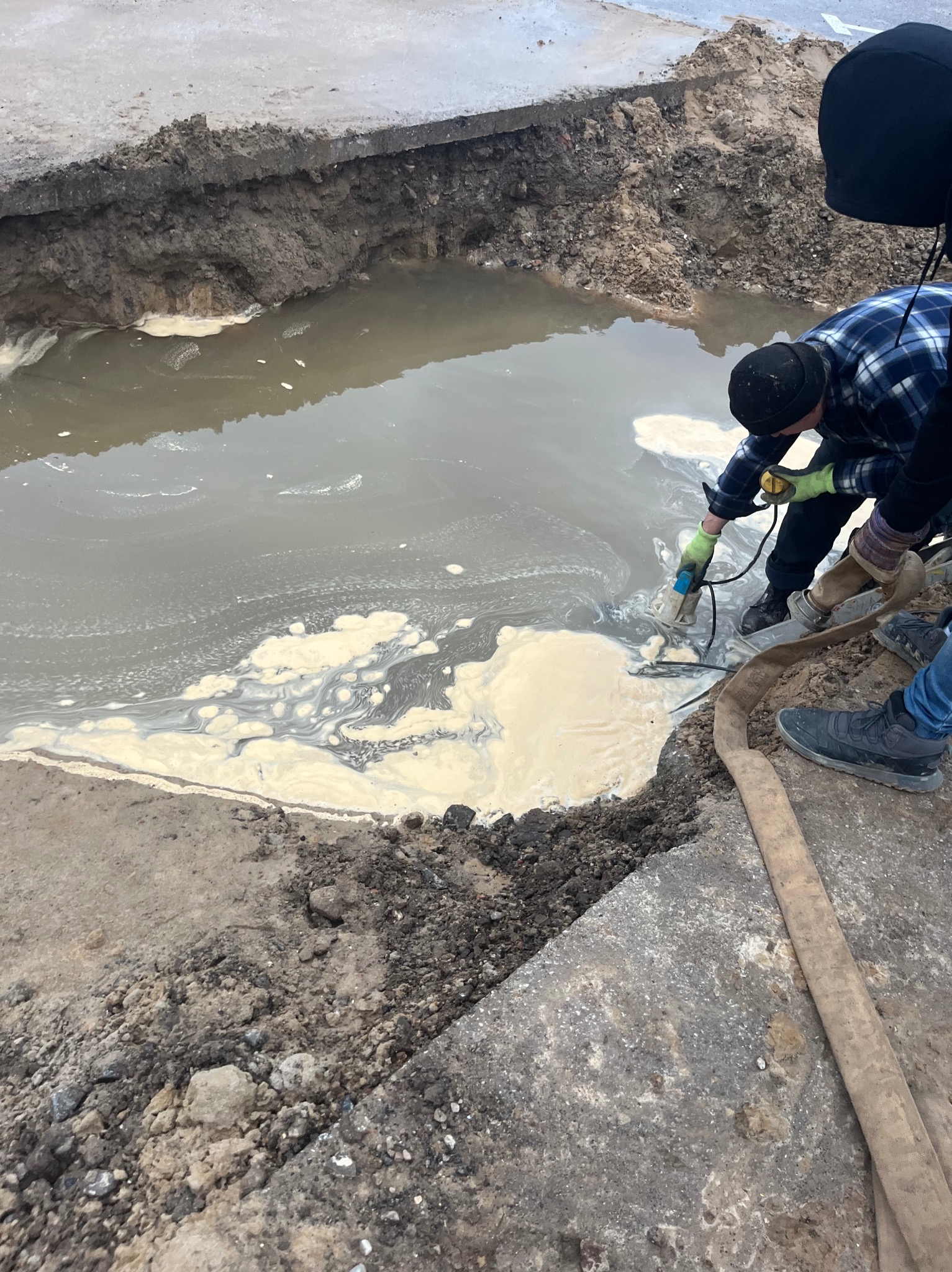 Pracownicy w trakcie czyszczenia wykopu wypełnionego wodą z białym osadem, używając pompy zanurzeniowej i węża. Widoczny fragment drogi z uszkodzoną nawierzchnią.