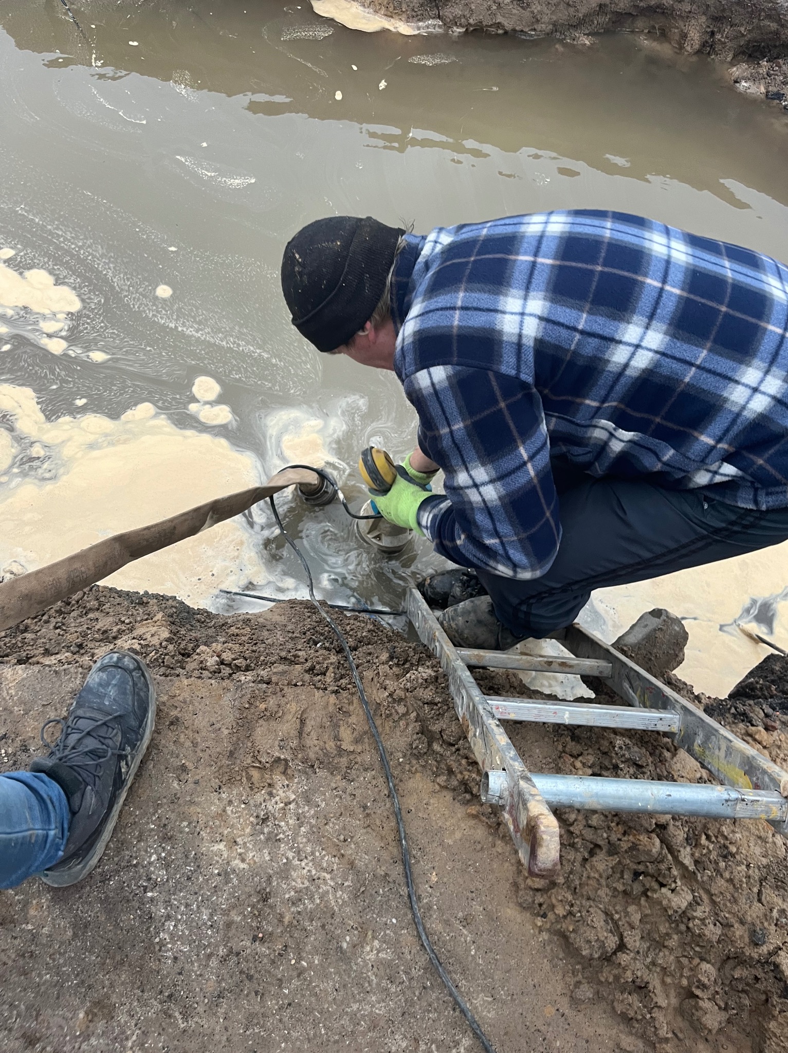 Pracownik w niebieskiej koszuli w kratę i czapce pracuje przy pompie zanurzonej w zbiorniku z brudną wodą, stojąc na aluminiowej drabinie.