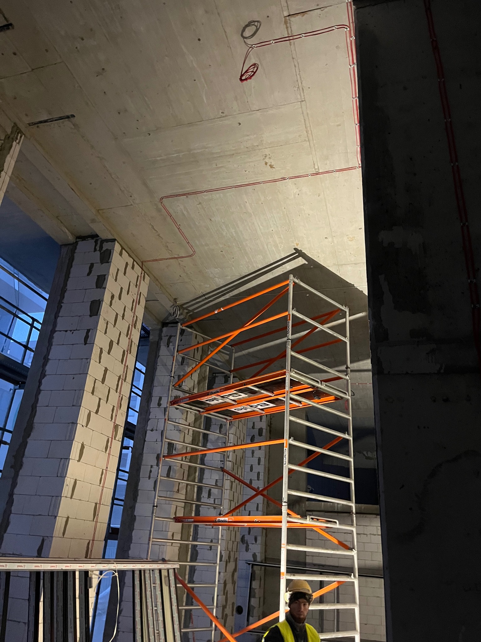 Wysokie wnętrze w surowym stanie z widocznymi instalacjami elektrycznymi na suficie i rusztowaniem, w tle ściana z bloków i okna, na pierwszym planie fragment sylwetki pracownika w kasku.