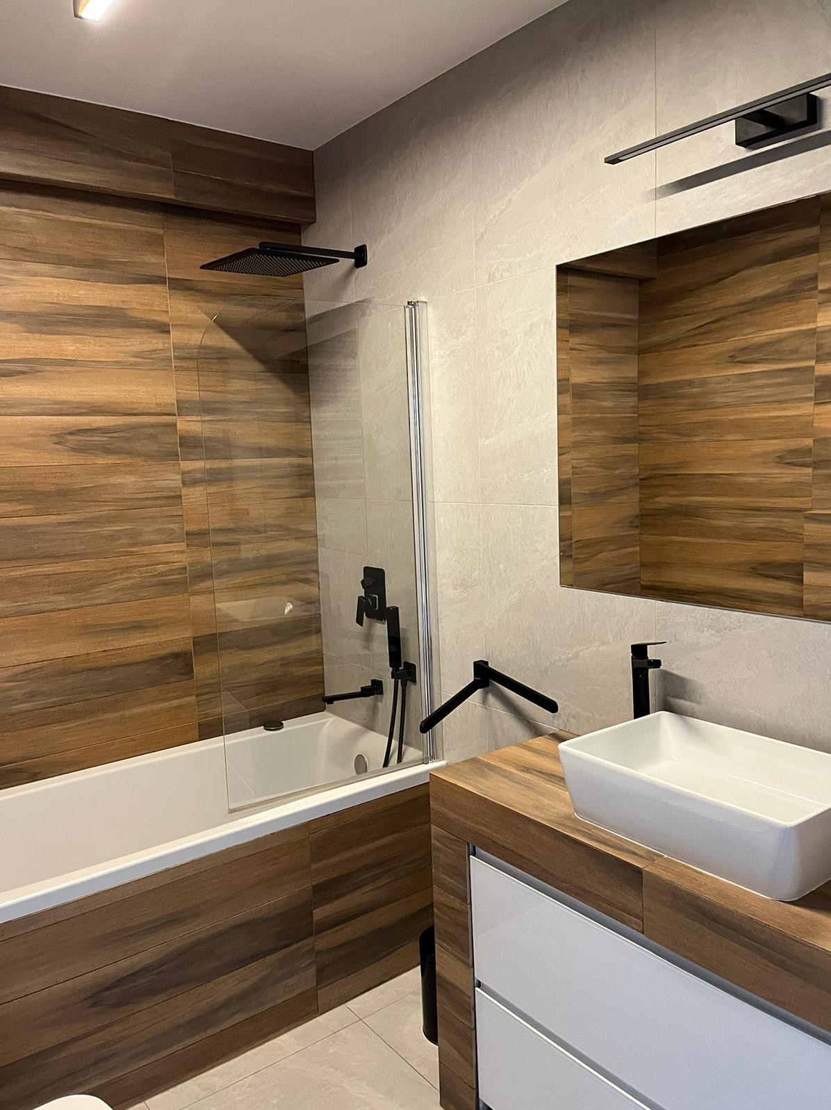 Nowoczesna łazienka z drewnopodobnymi płytkami na ścianach, białą wanną z czarną armaturą i umywalką nablatową na drewnianym blacie z białymi szufladami.