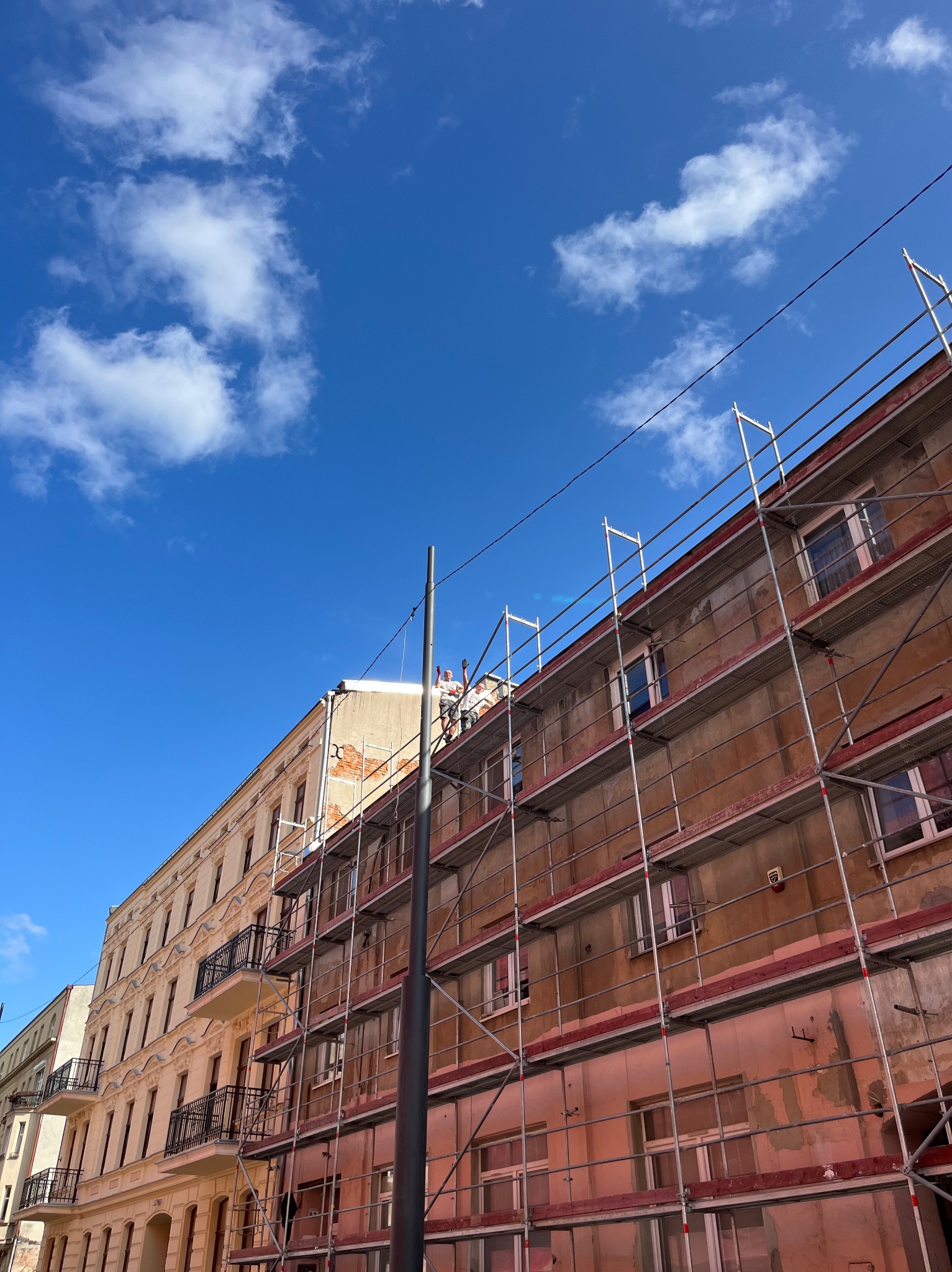 Budynek w trakcie renowacji elewacji z widocznymi rusztowaniami i pracownikami na dachu, na tle błękitnego nieba z chmurami.