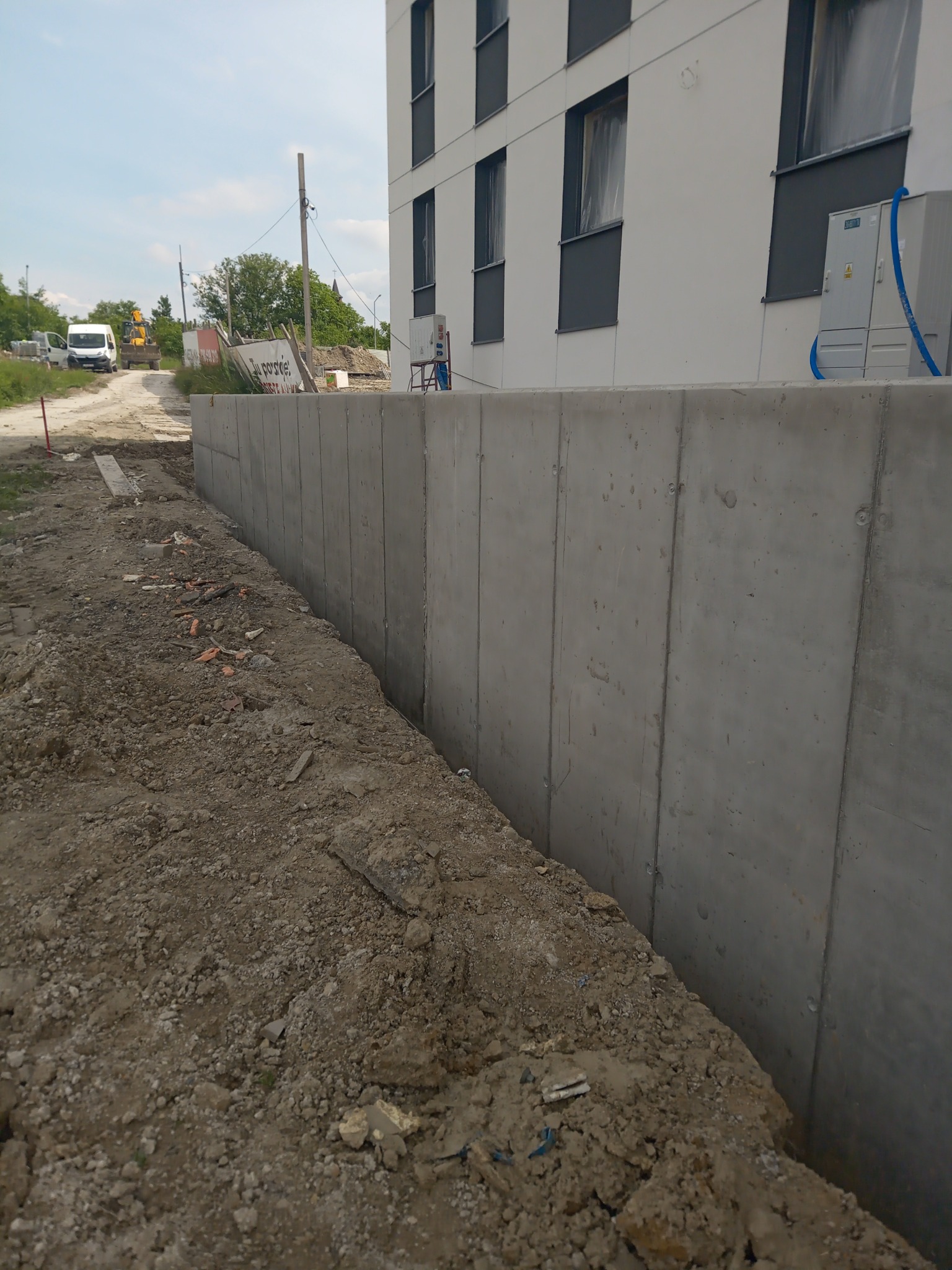 Wykop fundamentów pod budynek z widoczną ścianą fundamentową z betonu, na drugim planie budynek w trakcie budowy z zamontowanymi oknami i instalacją zewnętrzną.