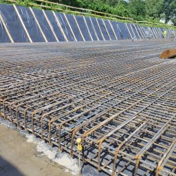 BartBud - Siatka zbrojeniowa fundamentu z żółtymi dystansami, widoczna folia izolacyjna i pracownicy w tle.