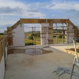 Wznoszenie ścian domu z betonu komórkowego, widoczne szalunki i rusztowania, w tle krajobraz wiejski pod błękitnym niebem.