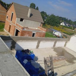 BartBud - Wznoszenie dwukondygnacyjnego domu z czerwonej cegły, widok z góry na budowę z widocznymi elementami konstrukcyjnymi i częściowo ocieplonym fundamentem, w tle krajobraz podmiejski z domami i zielenią.