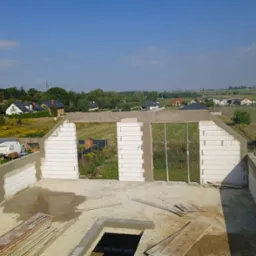 Widok z g&oacute;ry na budowę domu jednorodzinnego z białych bloczk&oacute;w, z widocznymi otworami okiennymi i zalanym betonem wieńcem, w tle krajobraz wiejski.