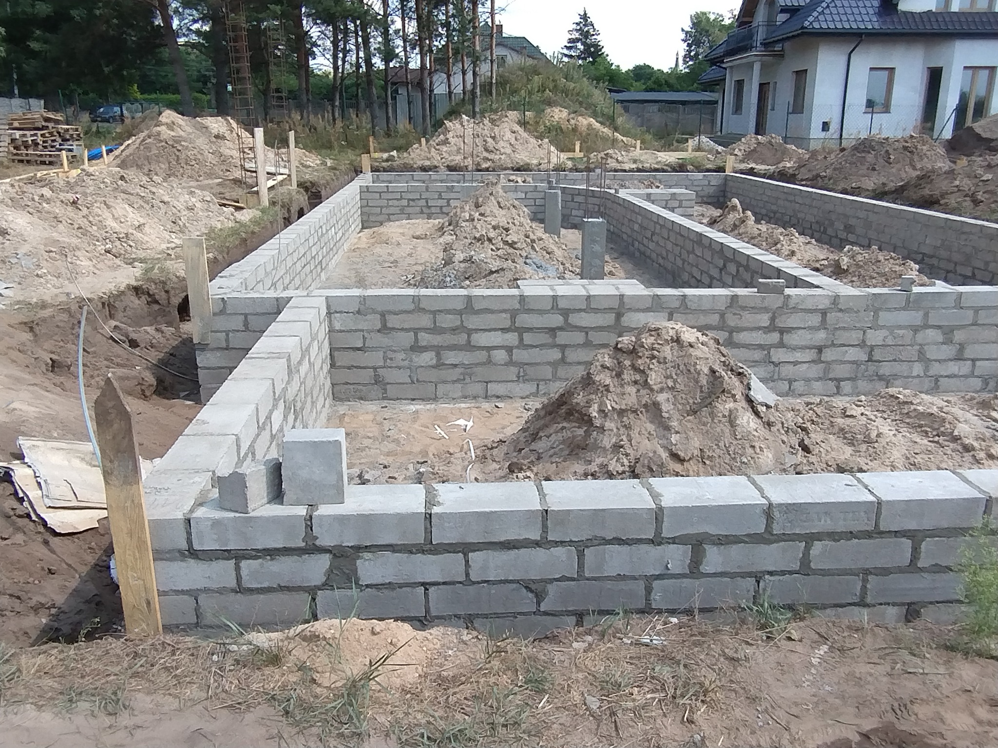 Wznoszenie muru fundamentowego z bloczków betonowych na placu budowy domu jednorodzinnego, widoczne wystające pręty zbrojeniowe i pryzmy ziemi.