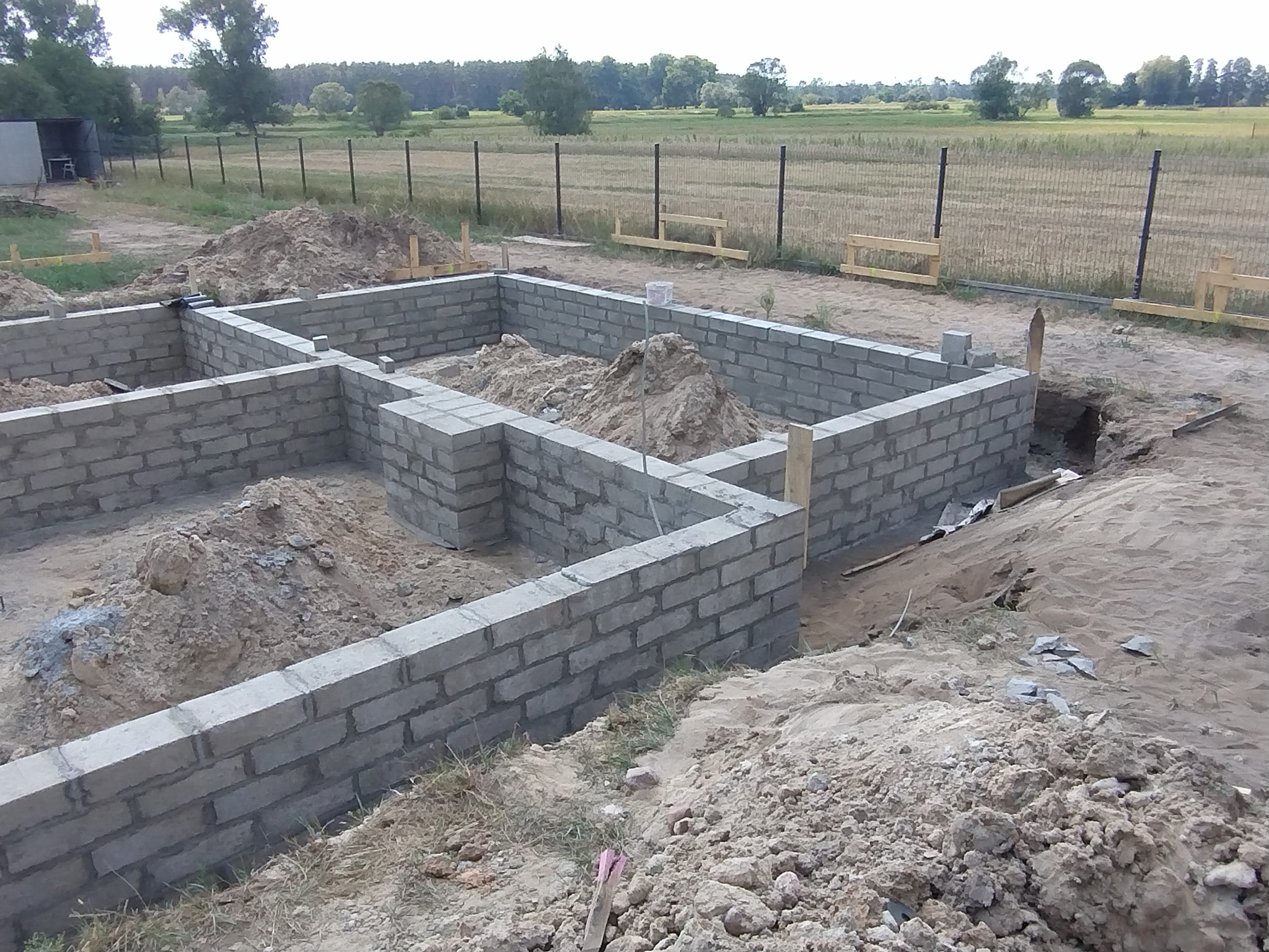 Świeżo wymurowane ściany fundamentowe z bloczków betonowych, częściowo zasypane ziemią, widok na teren budowy z ogrodzeniem i polem w tle.