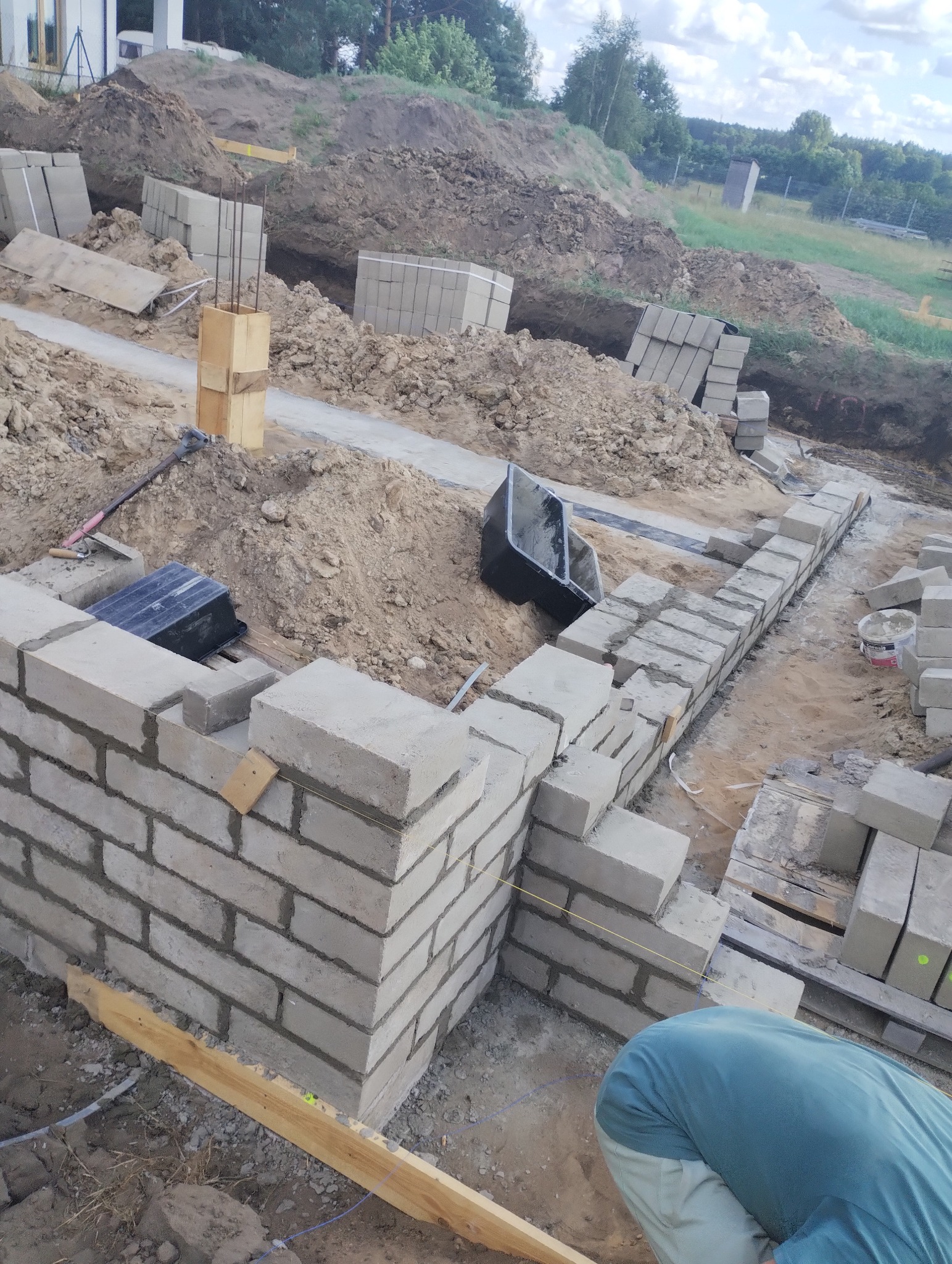 Wznoszenie muru z bloczków betonowych na fundamencie, widoczne narzędzia murarskie, sznurek traserski i zapas bloczków na palecie.