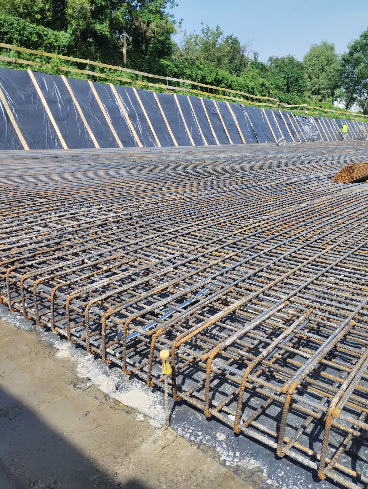 Siatka zbrojeniowa fundamentu z żółtymi dystansami, widoczna folia izolacyjna i pracownicy w tle.