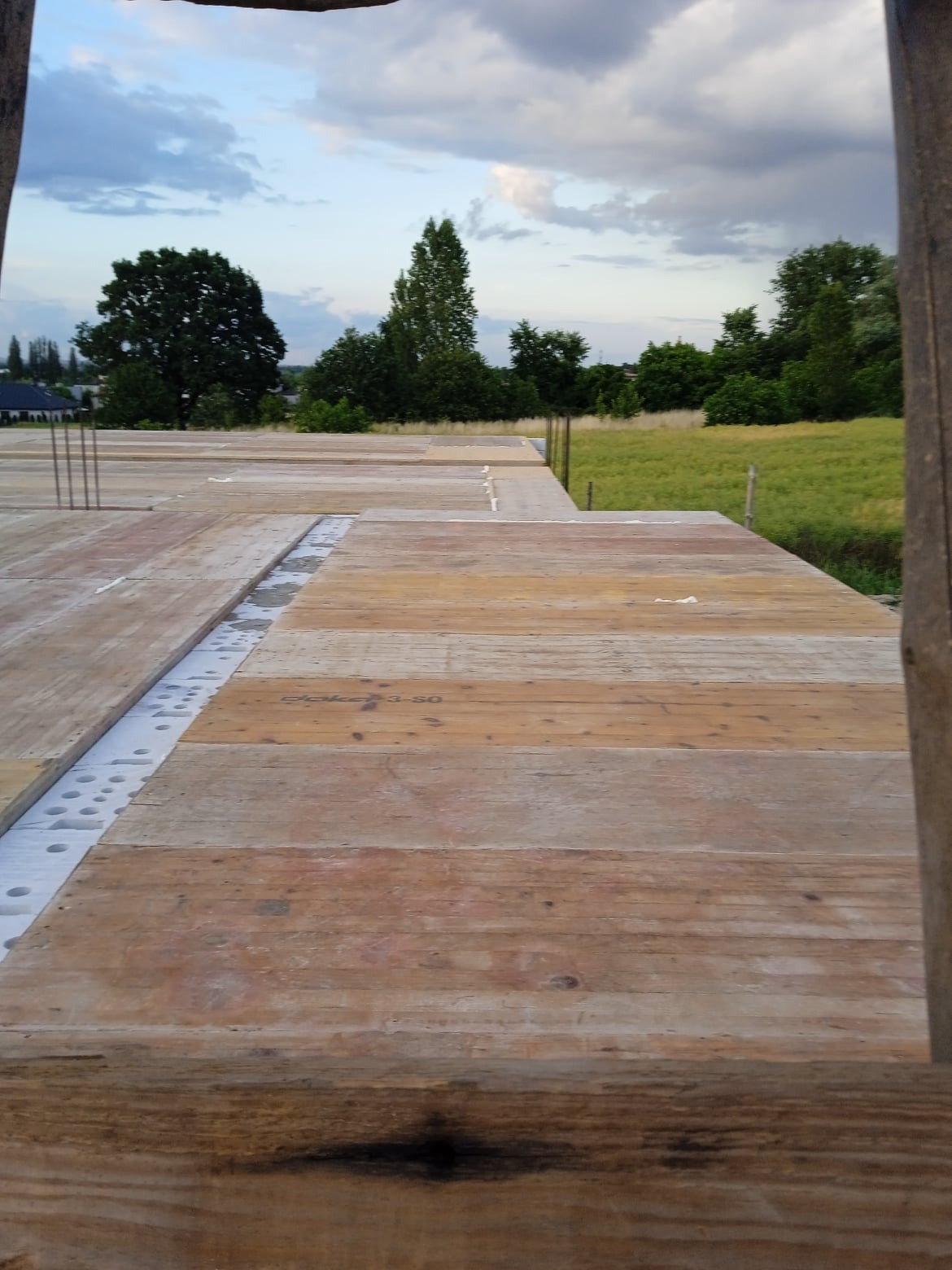 Deskowanie płyty fundamentowej z widoczną izolacją termiczną i wystającymi prętami zbrojeniowymi, na tle łąki i pochmurnego nieba.