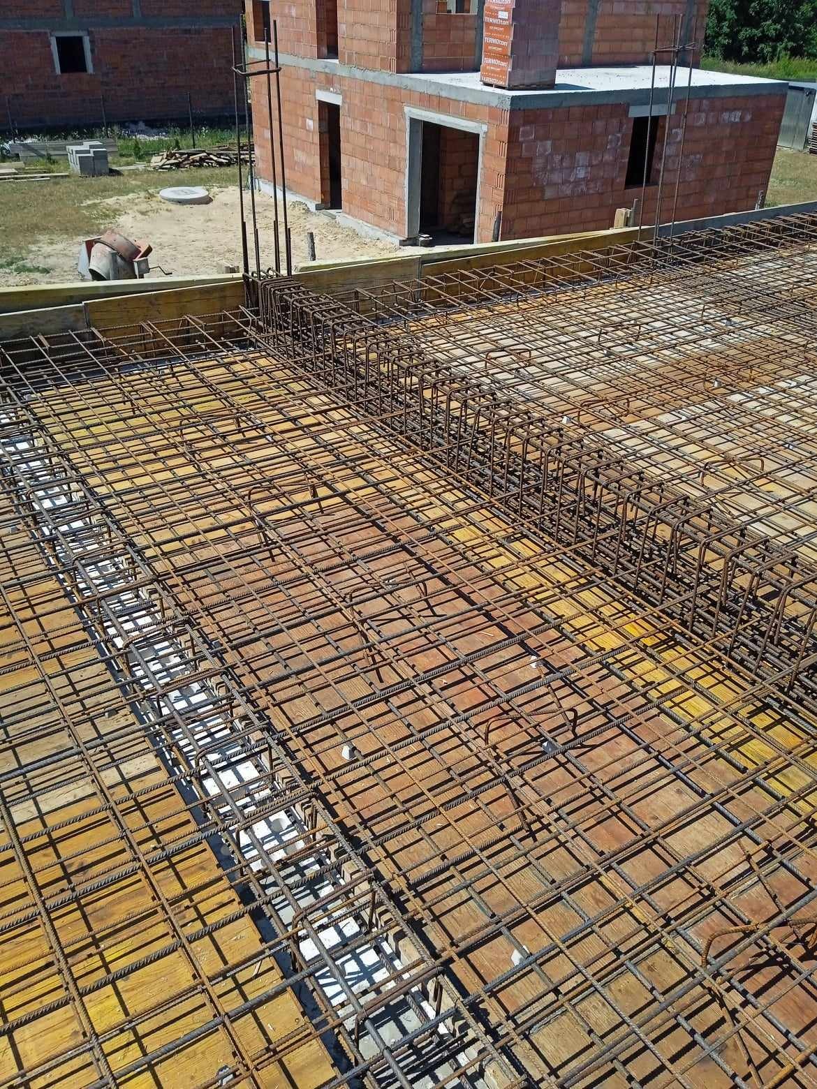 Zbrojenie płyty fundamentowej na deskowaniu, widoczny szkielet domu z czerwonej cegły w tle.