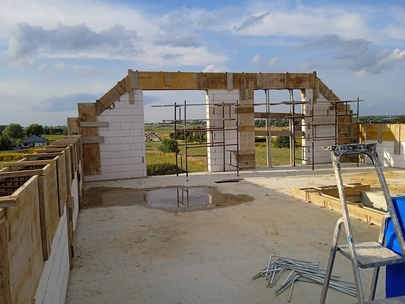 Wznoszenie ścian domu z betonu komórkowego, widoczne szalunki i rusztowania, w tle krajobraz wiejski pod błękitnym niebem.
