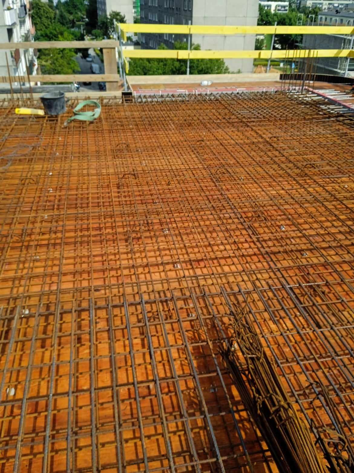 Siatka zbrojeniowa ułożona na deskowaniu, widok z góry, w tle budynki mieszkalne i elementy zabezpieczeń budowlanych.