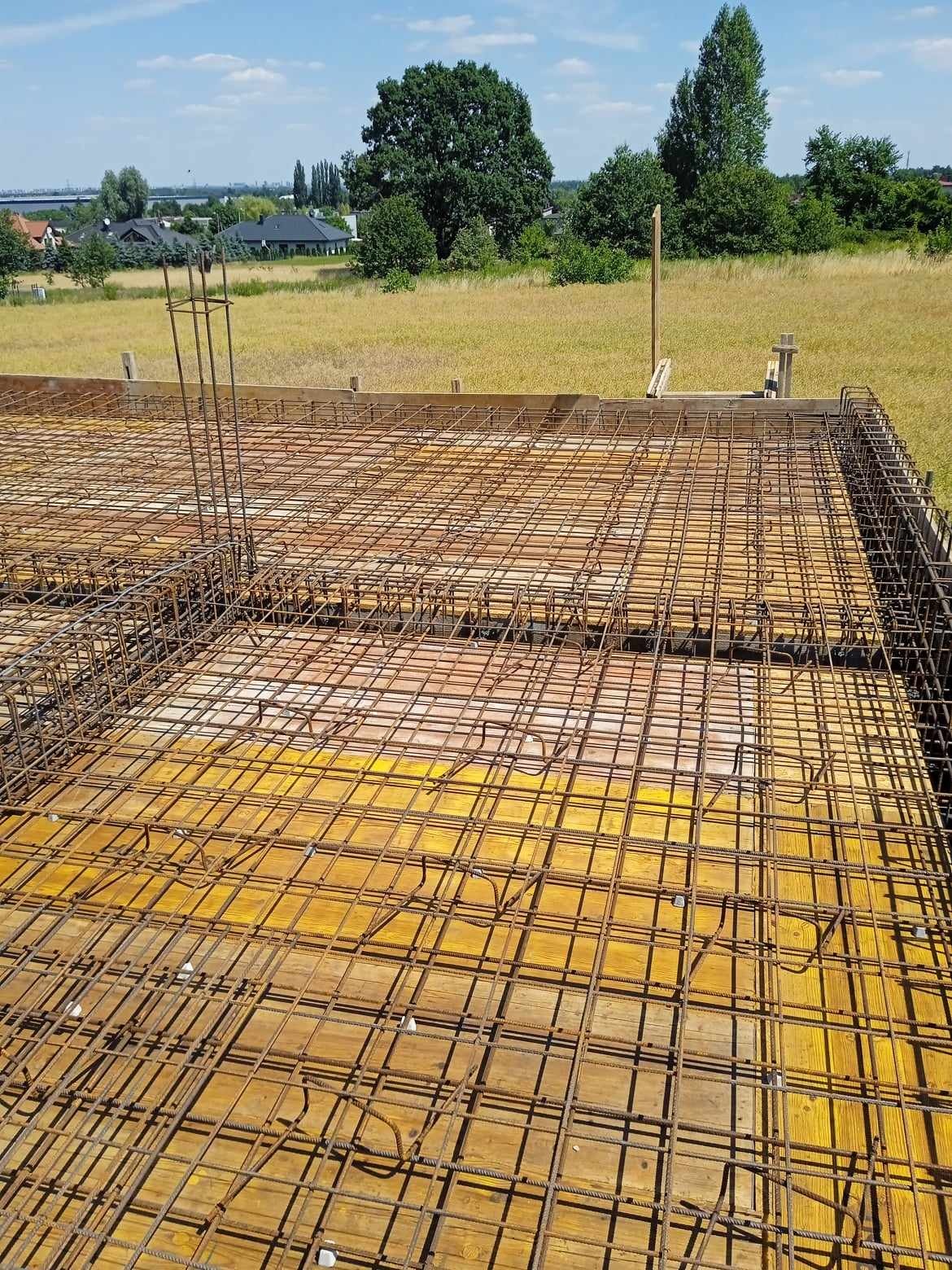 Zbrojenie płyty fundamentowej na deskowaniu z widocznymi wystającymi prętami pionowymi i otwartym krajobrazem w tle.